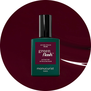 Un flacon de vernis à ongles Manucurist Paris Green Flash avec une étiquette vert foncé et un capuchon noir, sur un fond circulaire bordeaux profond.