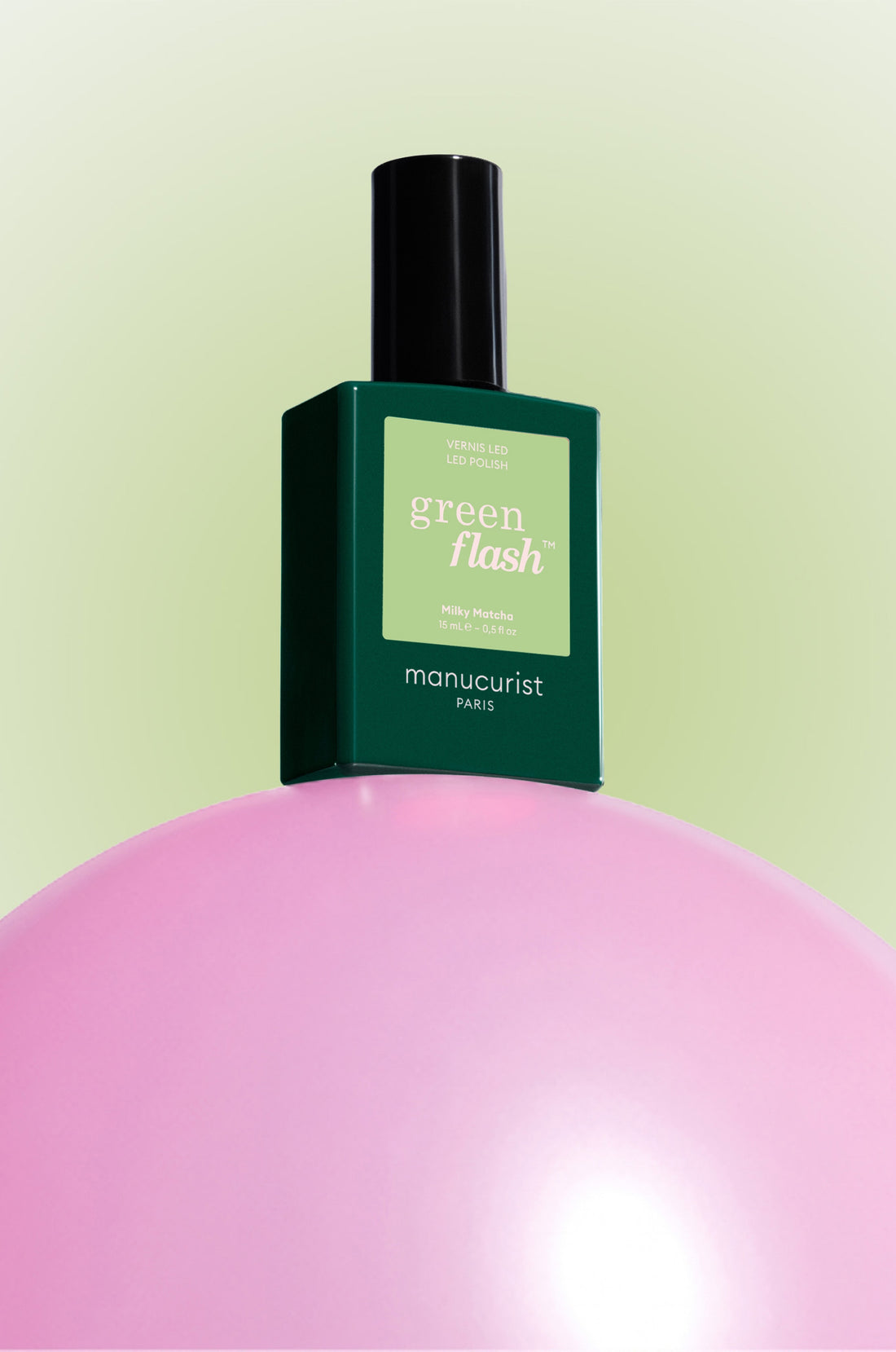A green bottle of Manucurist Green Flash nail polish is balanced on top of a glossy pink sphere, set against a light green gradient background.