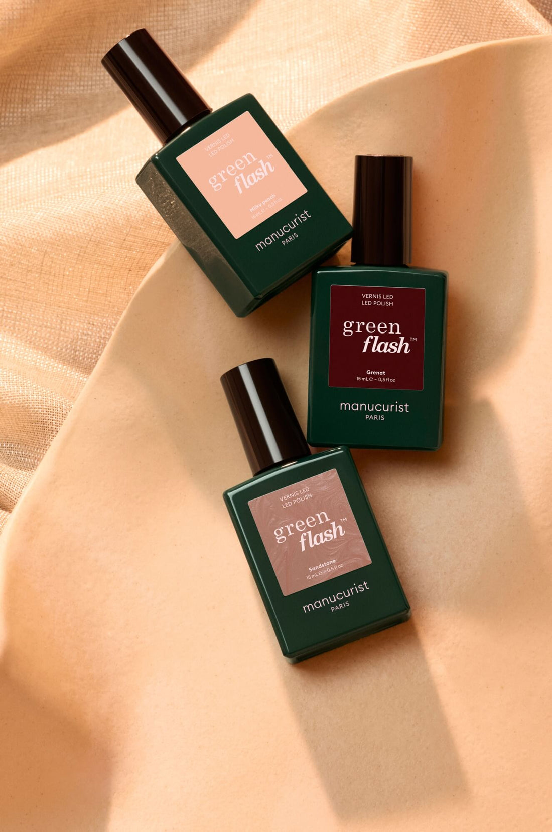 Three green bottles of Manucurist Green Flash nail polish are arranged on a beige surface with a soft, textured fabric background. Each bottle has a distinct label color and text.
