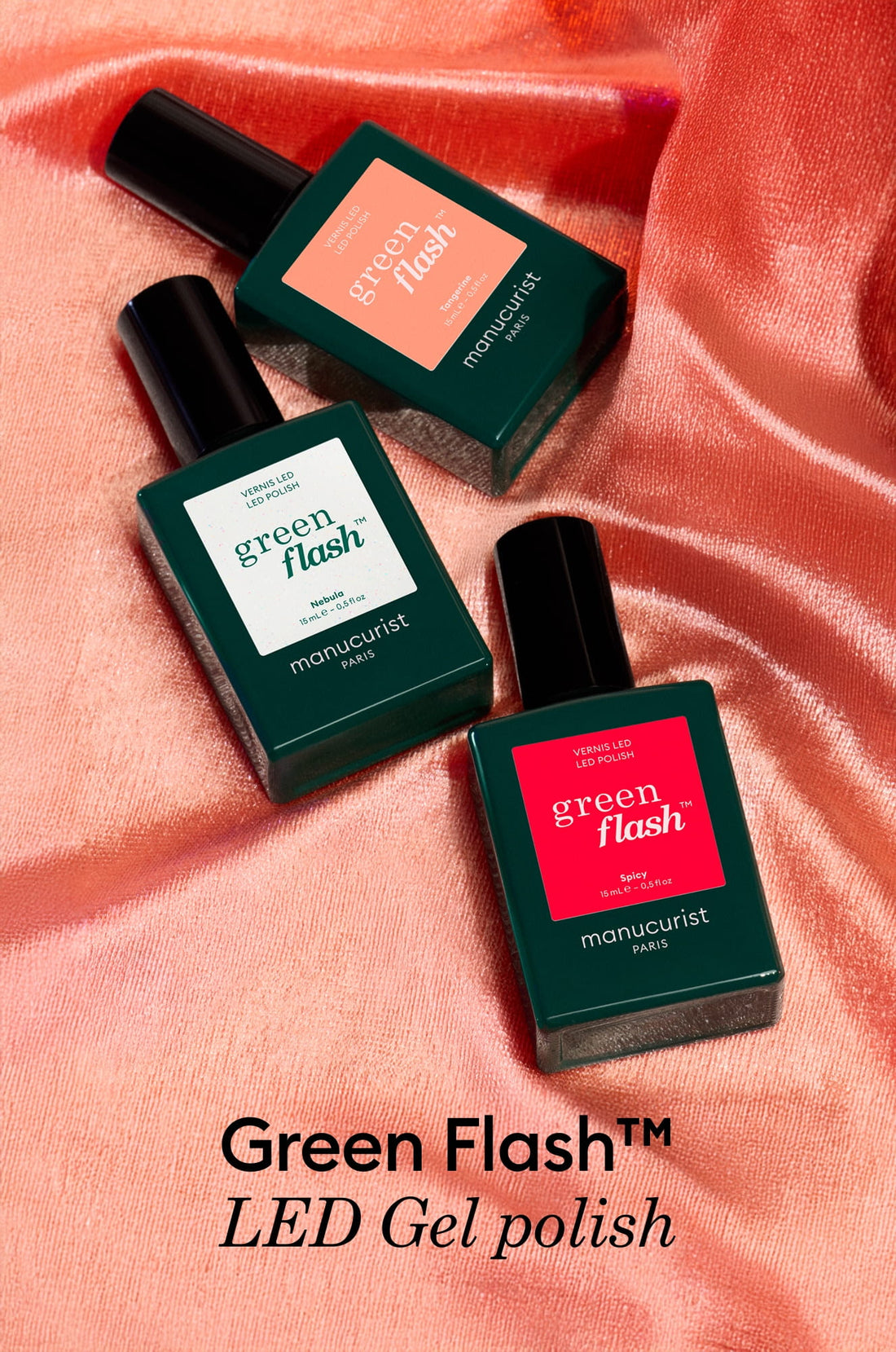 Three bottles of Green Flash LED gel polish by Manucurist Paris are arranged on shiny pink fabric. The bottles are dark green with different colored labels in coral, light green, and red.