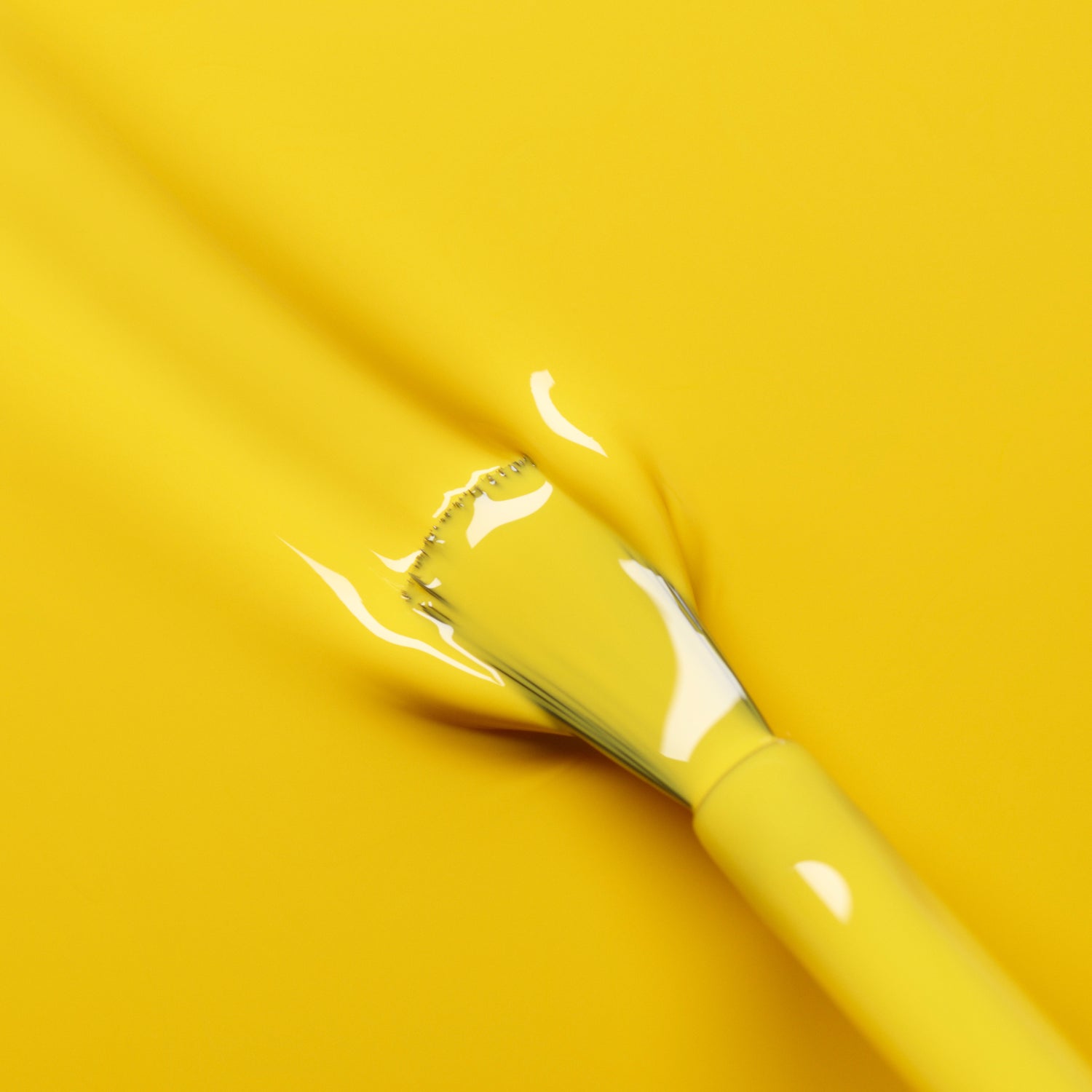 A paintbrush coated in Manucurist’s Gold Button nail polish glides smoothly over a yellow surface, blending perfectly into the background.