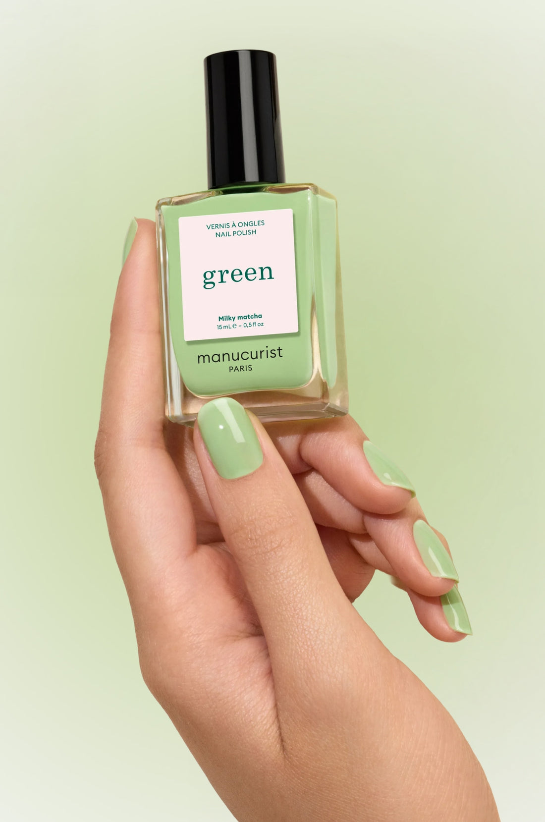A hand with light green painted nails holds a bottle of Manucurist Paris green nail polish in the same shade, labeled Milky Matcha. The background is a soft beige.