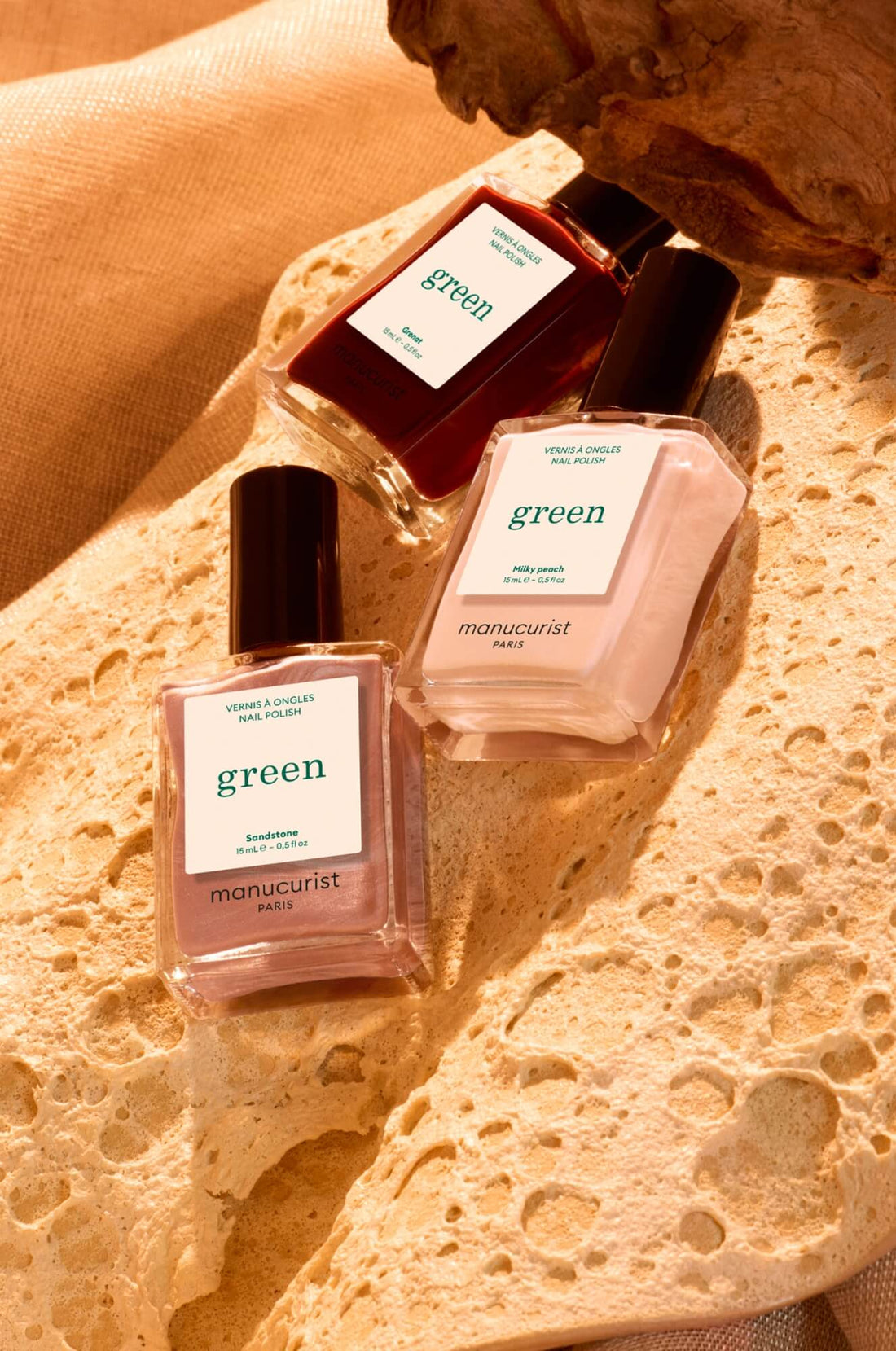Three bottles of Manucurist Paris Green nail polish in different shades are arranged on a textured, beige stone surface with warm lighting and a brown fabric background.