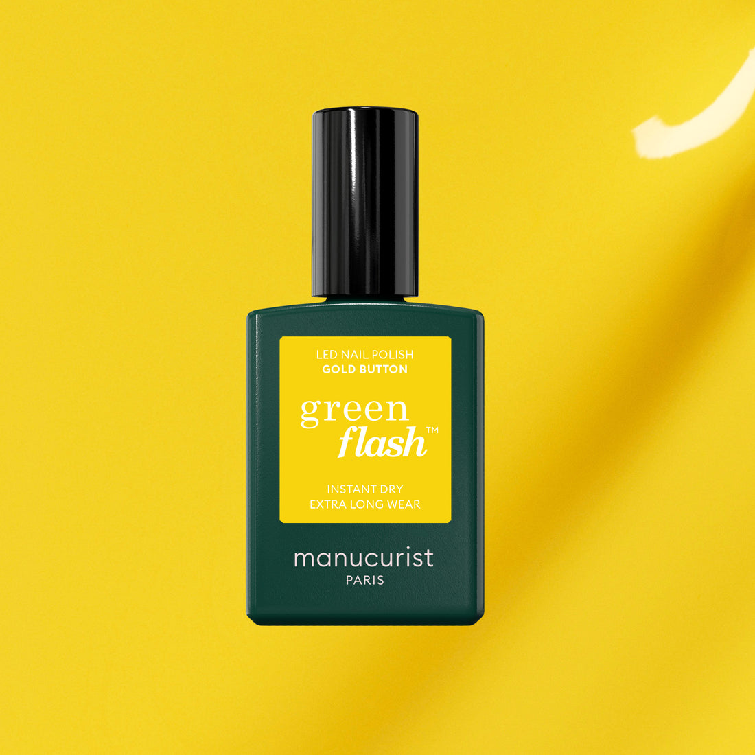 A bottle of Manucurist Gold Button gel polish with a black cap and green label, shown against a bright yellow background.