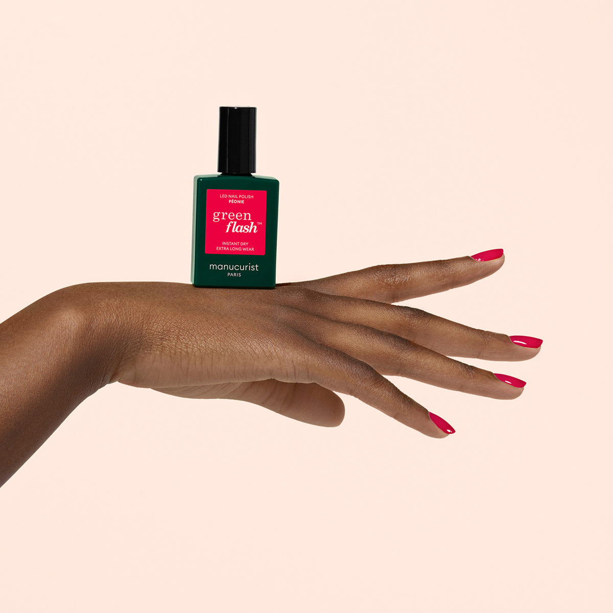 A hand with painted red nails is shown holding a green bottle of Manucurist Green Flash nail polish, balancing it on the back of the hand against a plain beige background.