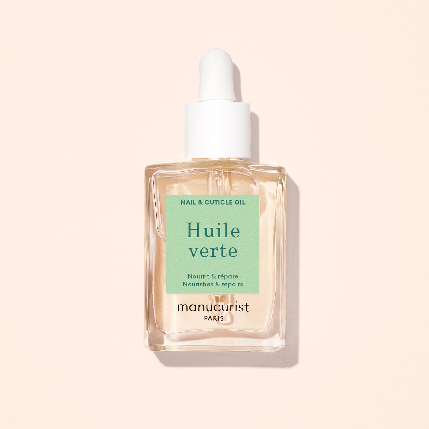 A clear dropper bottle of Manucurist Nourishing Cuticle Oil rests on a light beige background, featuring a green label with white and black text.