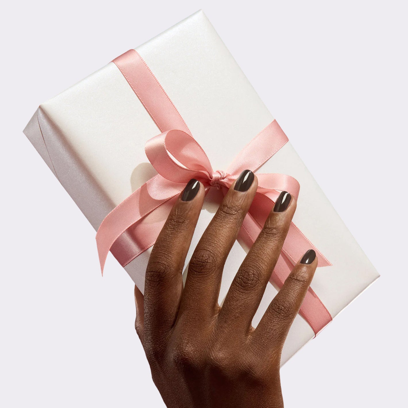 A hand with dark nail polish holds a white gift box wrapped with a pink ribbon and bow against a plain light background.