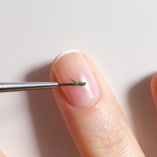 A close-up of a finger with a French manicure. A thin brush is painting a small green leaf design on the nail above the white tip. The background is plain and light.