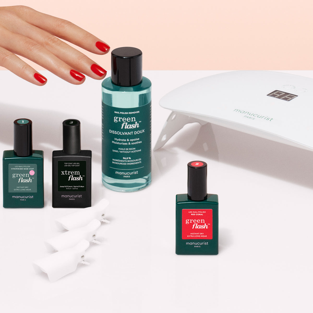 Une main aux ongles rouges se tend vers le Discovery Kit Manucurist, comprenant un dissolvant, un vernis gel Green Flash, des clips à ongles et une lampe LED sur une surface blanche.