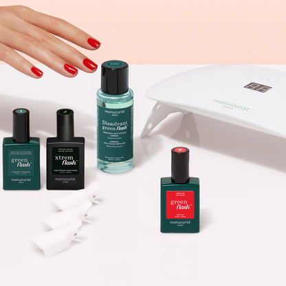 A hand with red nails is next to the Manucurist Discovery Kit, which includes nail polish, a vitaminized base coat, remover, a white LED lamp, and nail clips—everything needed for an ultra-glossy manicure.