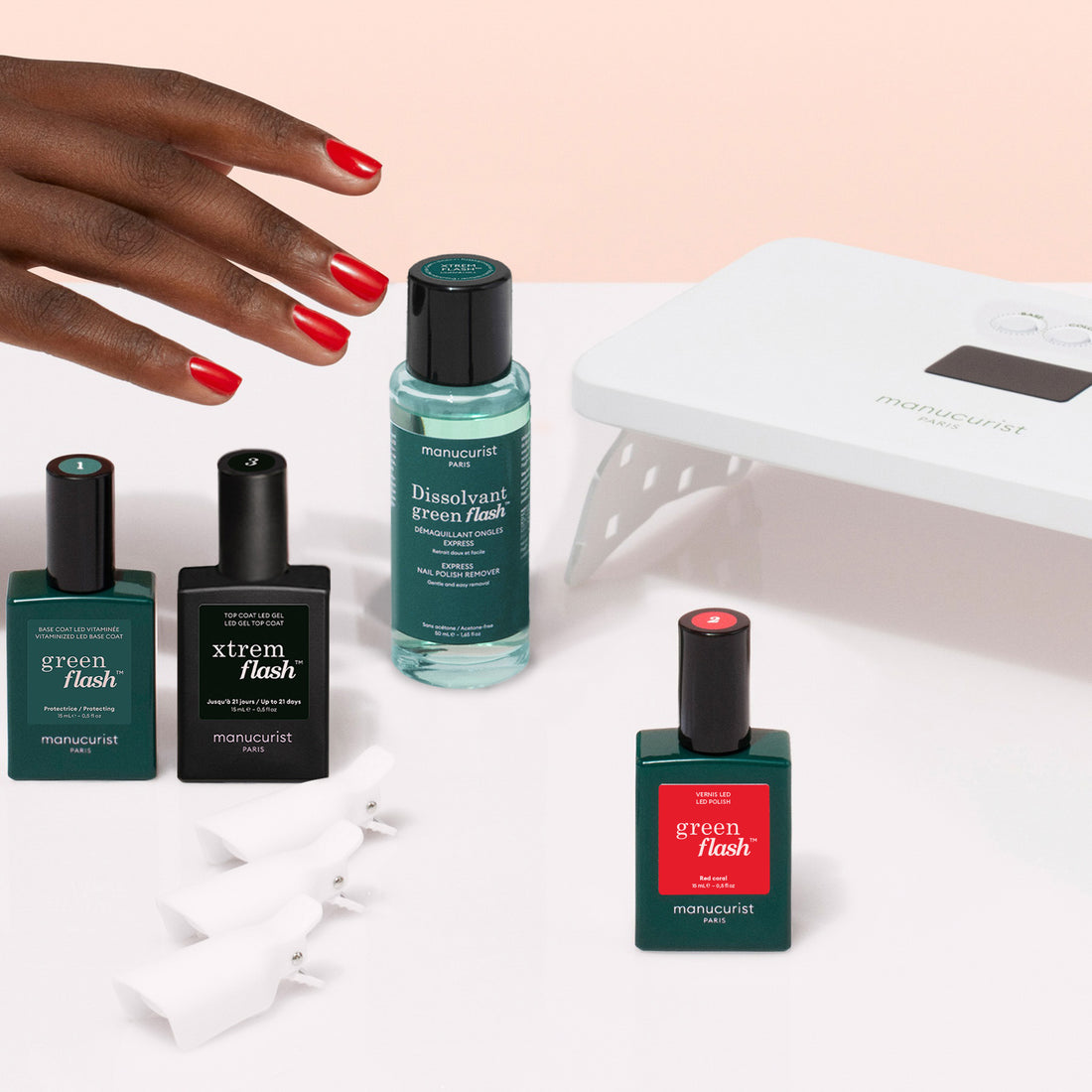A hand with red-painted nails reaches for the Manucurist Discovery Kit—featuring nail polish, a vitaminized base coat, a white LED nail lamp, and white clips—essentials for an ultra-glossy manicure on a light surface.