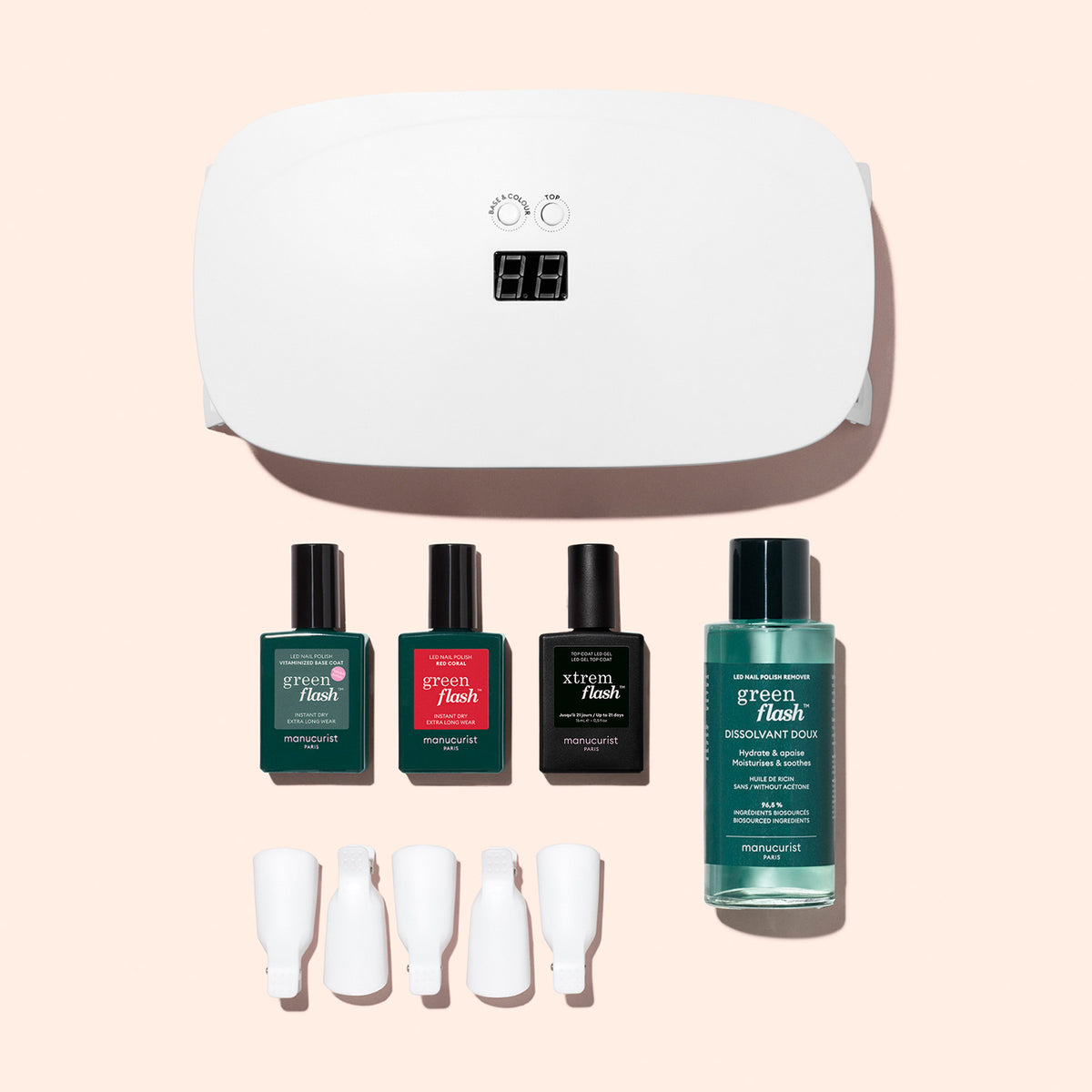 Le coffret découverte Manucurist, comprenant une lampe LED blanche pour ongles, trois vernis gel Green Flash, un dissolvant et quatre pinces blanches, est présenté sur un fond rose clair.