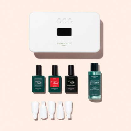 The Manucurist Discovery Kit features a white LED lamp, three nail polishes, a vitaminized base coat, remover, and four white nail clips—perfect for an ultra-glossy manicure—beautifully displayed on a light pink background.