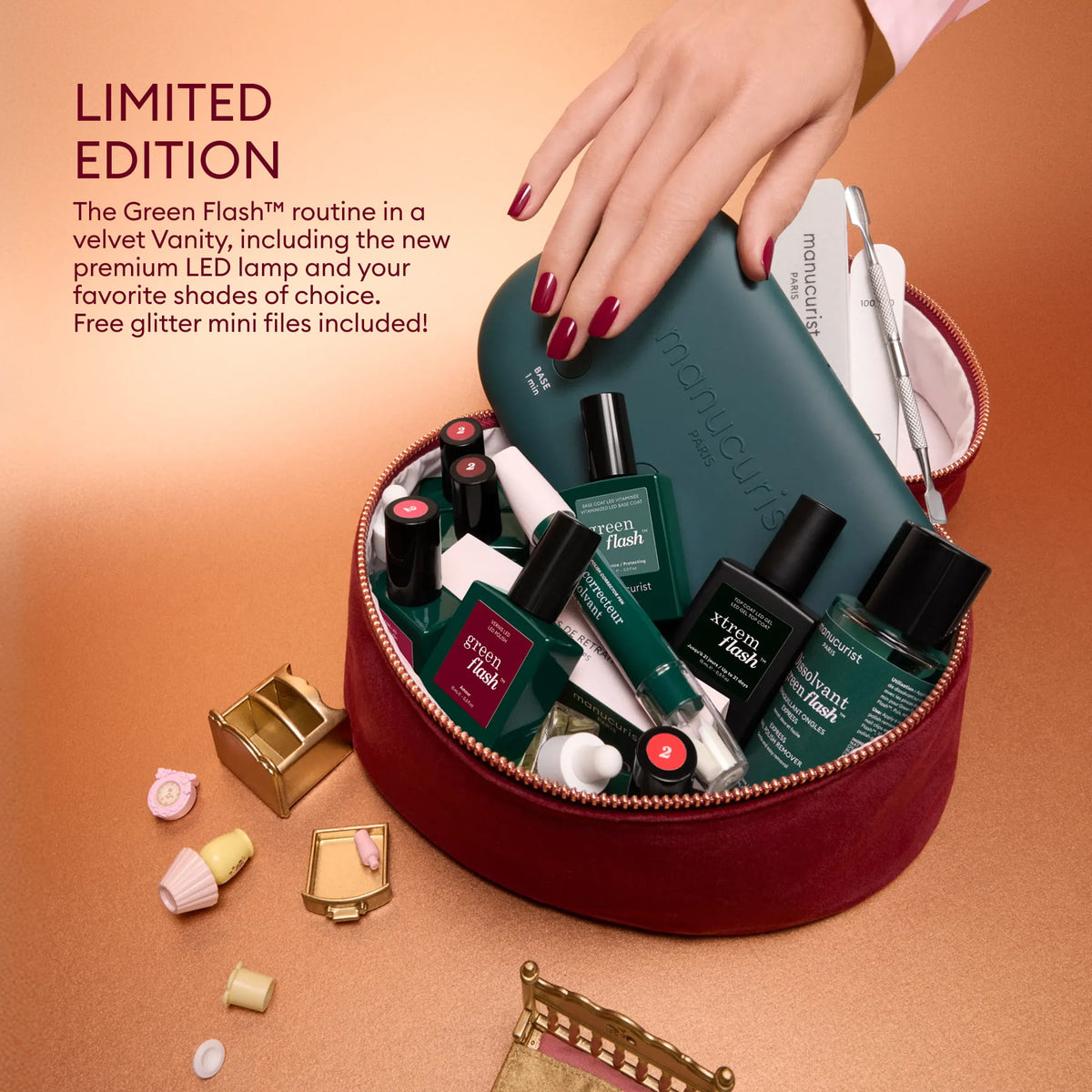 A hand holds the Manucurist Green Flash™ Discovery Kit Gift Set, featuring nail essentials like polish bottles, a green LED lamp for salon-quality manicures at home, and mini nail files—all nestled inside a round velvet pouch marked “LIMITED EDITION.” Small decorative items are scattered around the set.