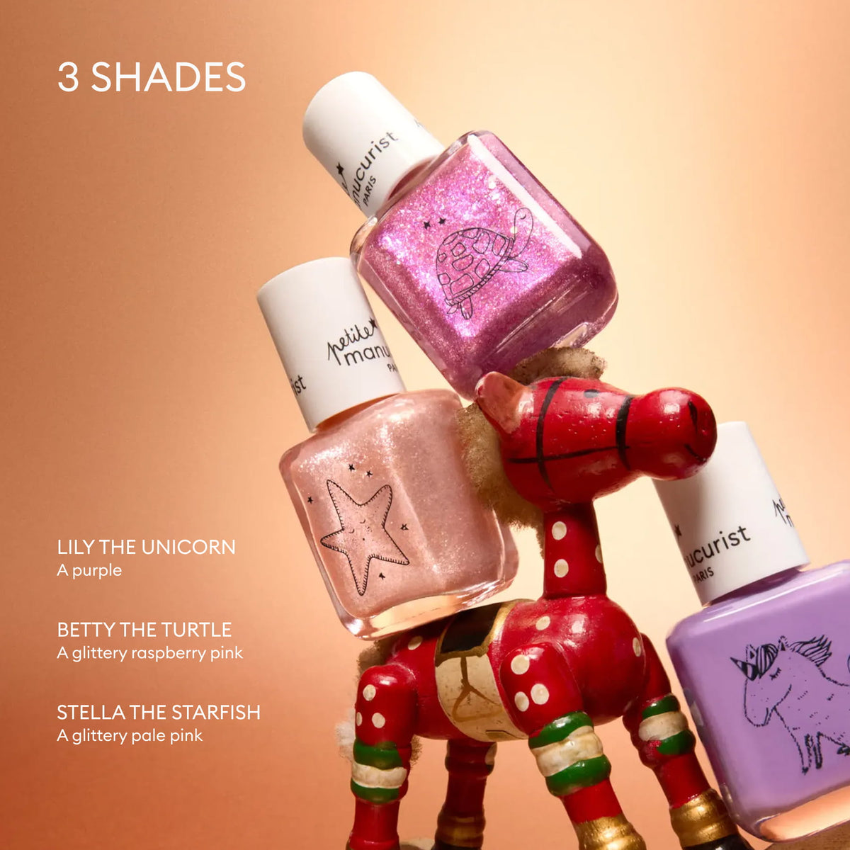 Three bottles from the Manucurist Petite Manucurist Trio—children’s nail polish in purple, glittery raspberry pink, and glittery pale pink—are stacked on a wooden toy reindeer against a peach background. Made with natural origin ingredients, each bottle features an adorable animal illustration, making them the perfect holiday gift.