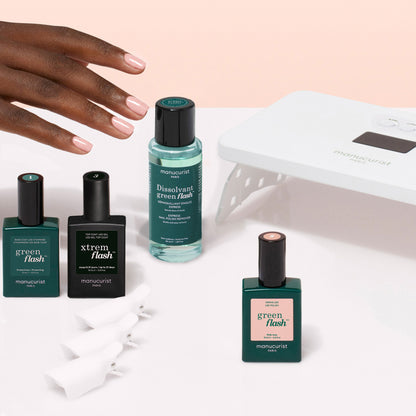 A hand with manicured pink nails rests above a Manucurist Discovery Kit - Soap Nails LED Gel Polish, which includes three bottles of polish, a remover, a white LED lamp, and plastic nail clips for an ultra-glossy manicure.