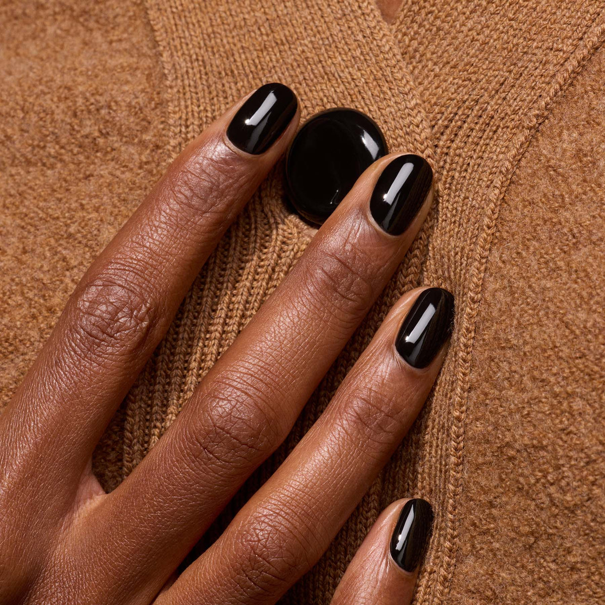 Une main portant le vernis à ongles noir Licorice de Manucurist posée sur un pull texturé marron près d&