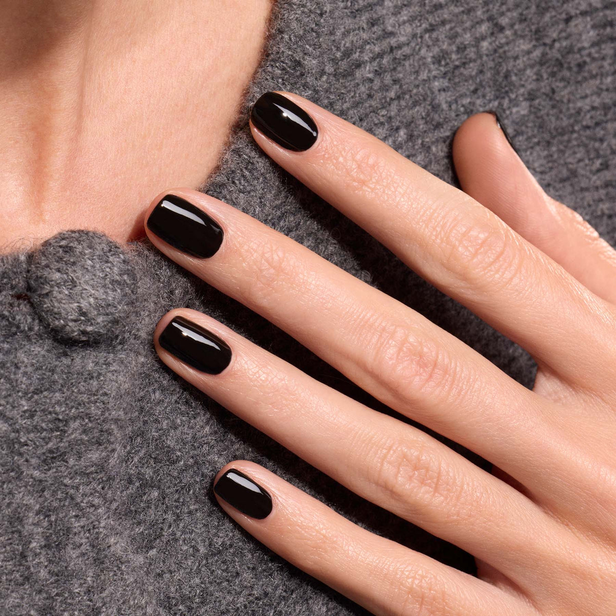 Une main aux ongles impeccablement manucurés avec le vernis Licorice de Manucurist repose sur un manteau texturé gris, orné d&
