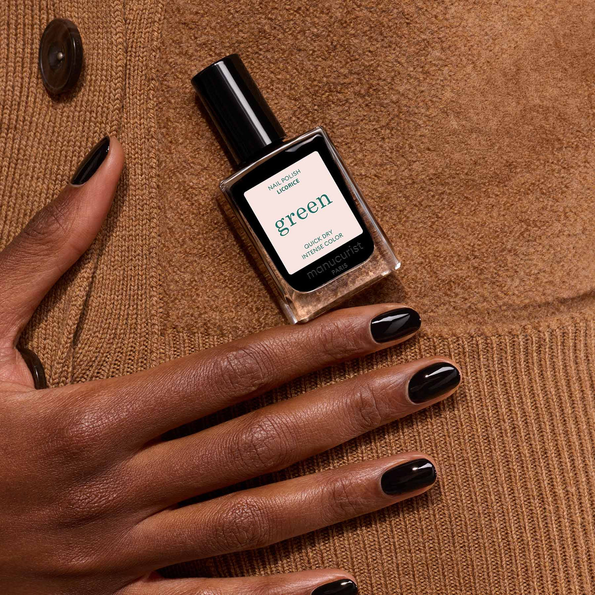 Une main aux ongles vernis noirs, chics et brillants repose sur du daim marron, tenant un flacon de vernis à ongles Manucurist Licorice.