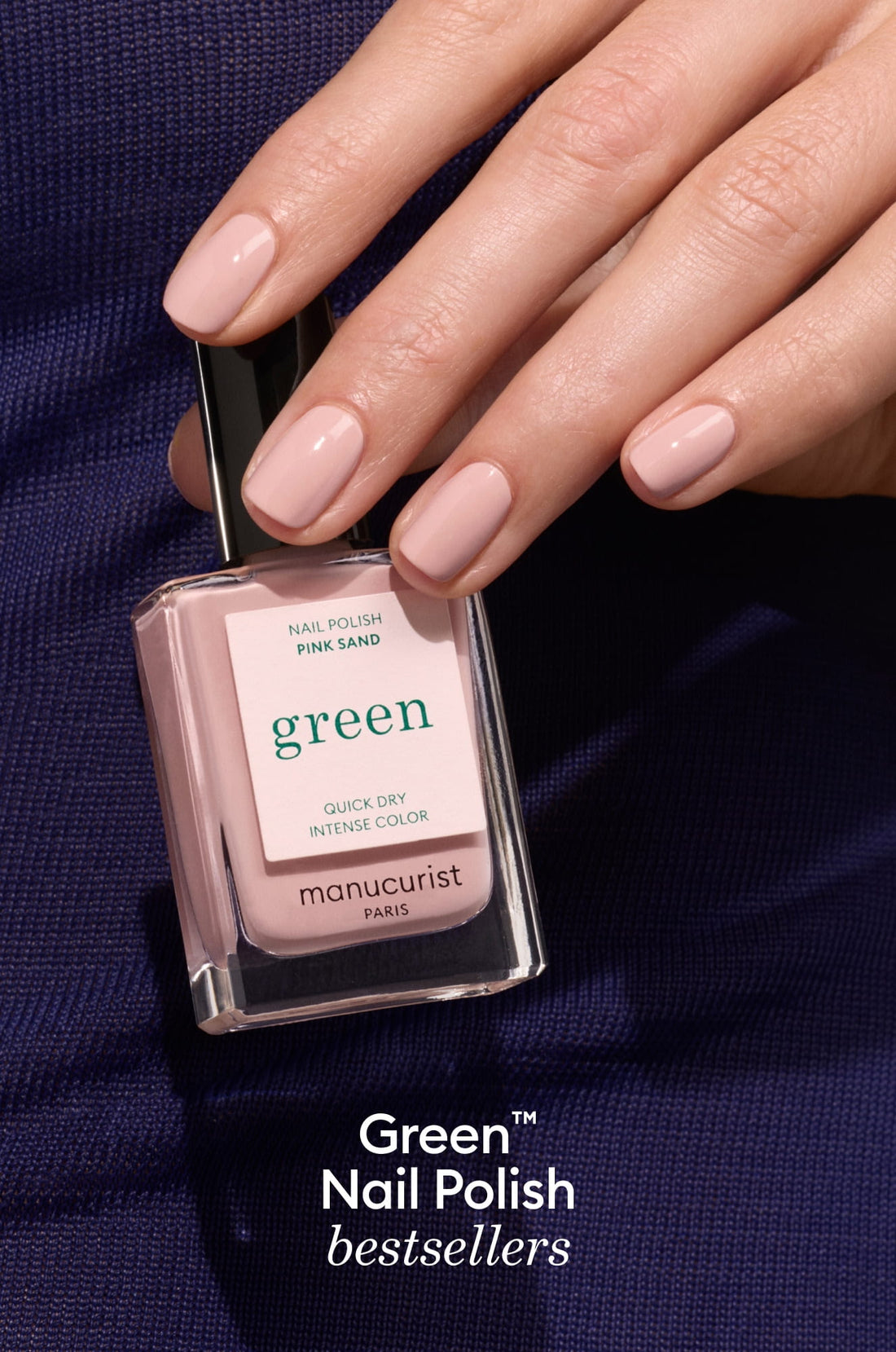 A hand with neatly manicured, light pink nails holds a bottle of Manucurist Paris Green nail polish in the shade Pink Sand against a dark fabric background. The text reads: Green Nail Polish bestsellers.