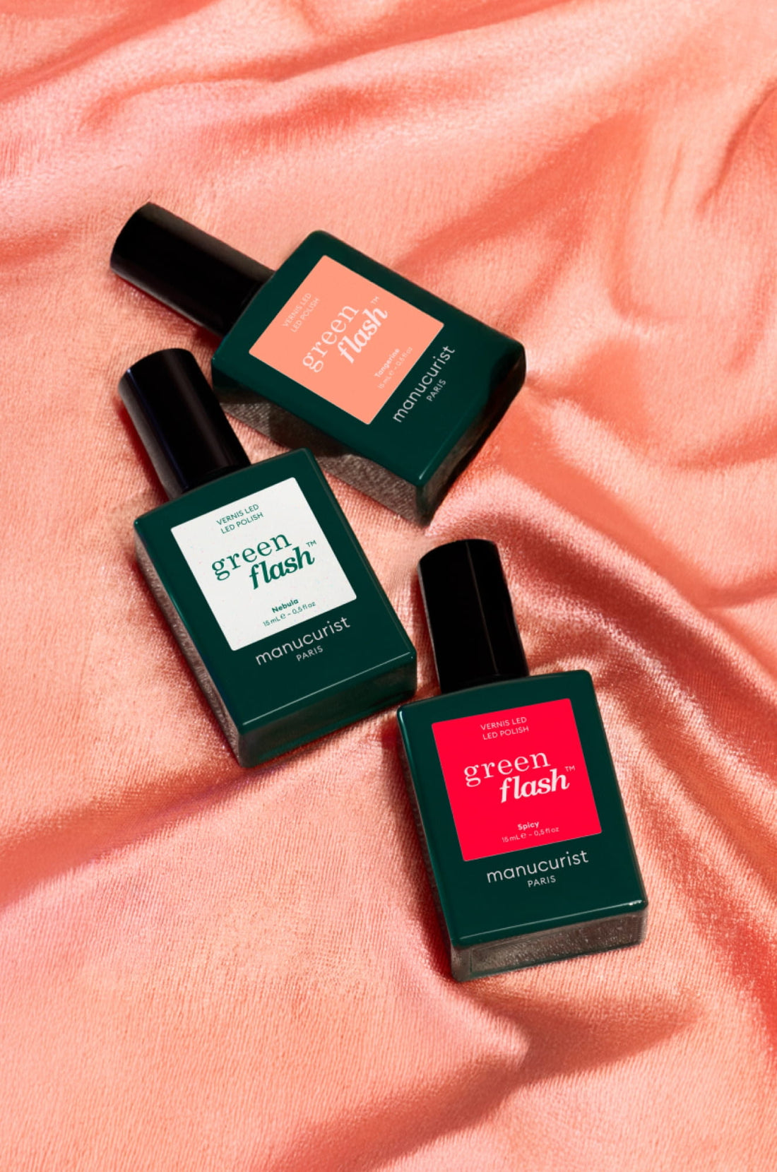 Three bottles of Manucurist Paris Green Flash nail polish in green square bottles with different colored labels, placed on a shiny pink satin fabric background.