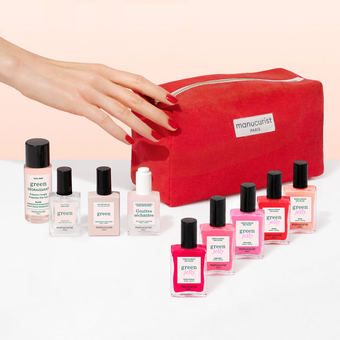 A hand with red nails reaches for a red Manucurist Super Starter Kit - Jelly Nail Polish manicure bag, surrounded by bottles of Manucurist plant-based nail polish in various colors and nail care products, all arranged on a white surface.
