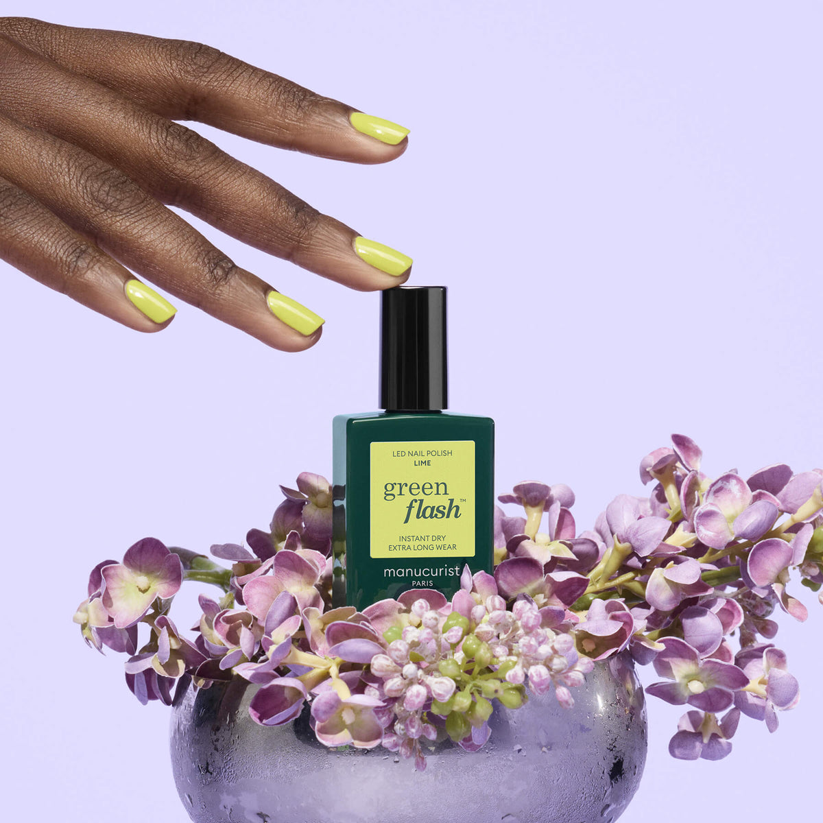 Une main aux ongles peints en jaune-vert survole un flacon de vernis gel Manucurist Lime chartreuse, entouré de fleurs violettes dans un vase transparent, sur fond lavande pâle.