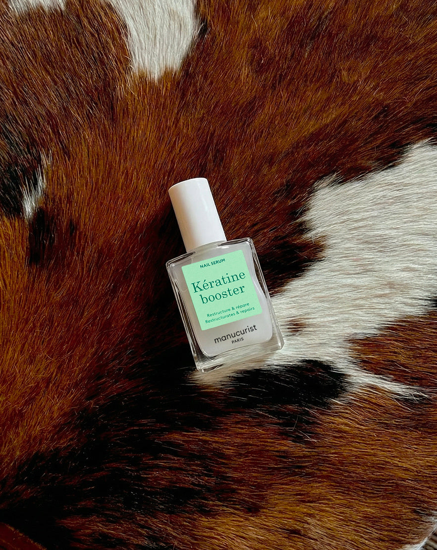 A bottle of Manucurist Kératine Booster nail serum with a white cap lies on a brown and white cowhide surface.