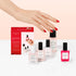 A hand with red-painted nails reaches for the Manucurist Discovery Kit - Jelly Nail Polish, featuring plant-based nail polish and care items in a red box, all displayed on a white surface with a light pink background.