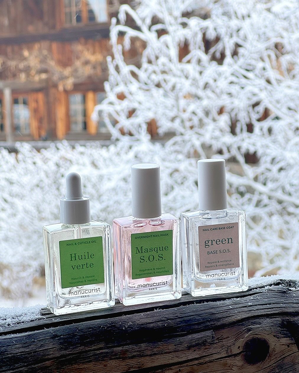 Three bottles of Manucurist nail care products are lined up on a wooden ledge, with a snowy, frost-covered tree and rustic wooden building in the background.