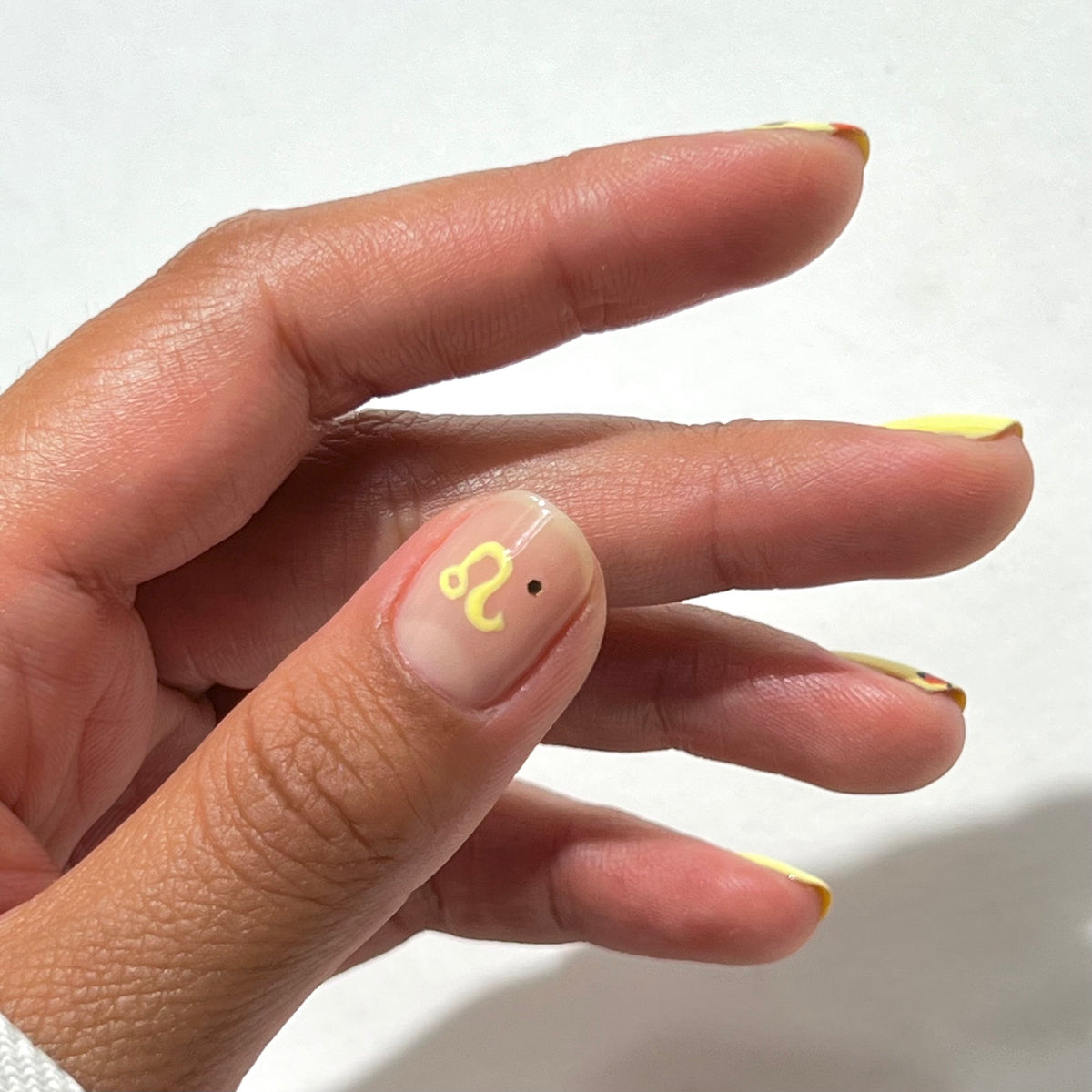 A hand with nails painted with yellow tips; the thumbnail features the Leo zodiac symbol in yellow and a small black dot on a clear base.
