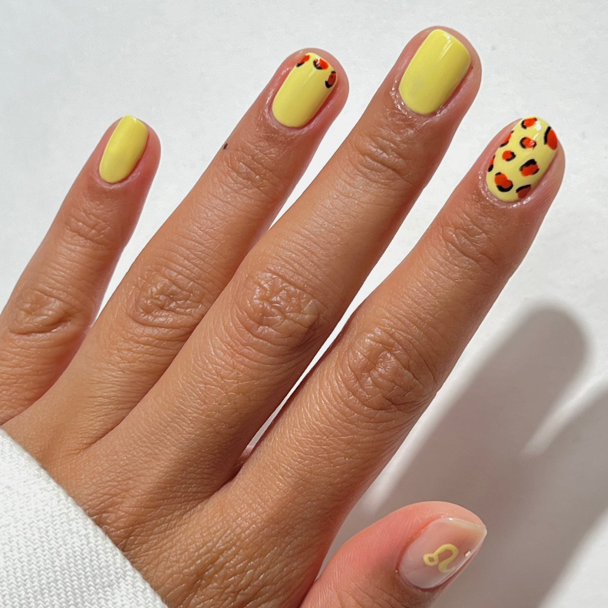 A hand with short, almond-shaped nails painted yellow, featuring two nails with orange and black cheetah print designs, and the pinky nail with a small yellow zodiac Leo symbol on a clear base.