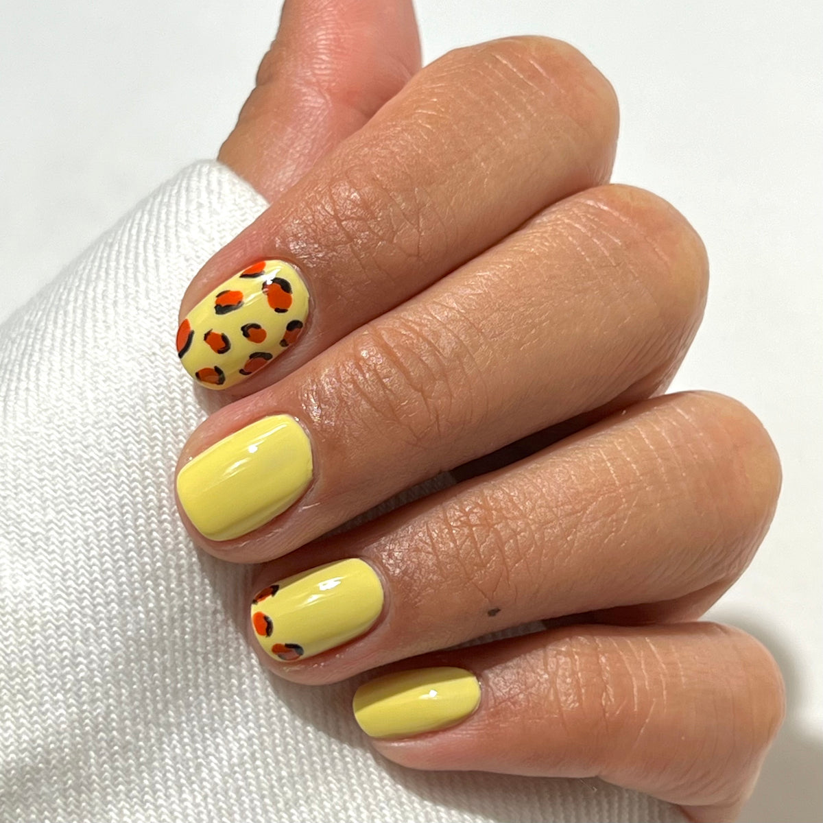 A hand with short, almond-shaped nails painted yellow; two nails feature a leopard print design in black and orange on a yellow base. The hand rests on white fabric.