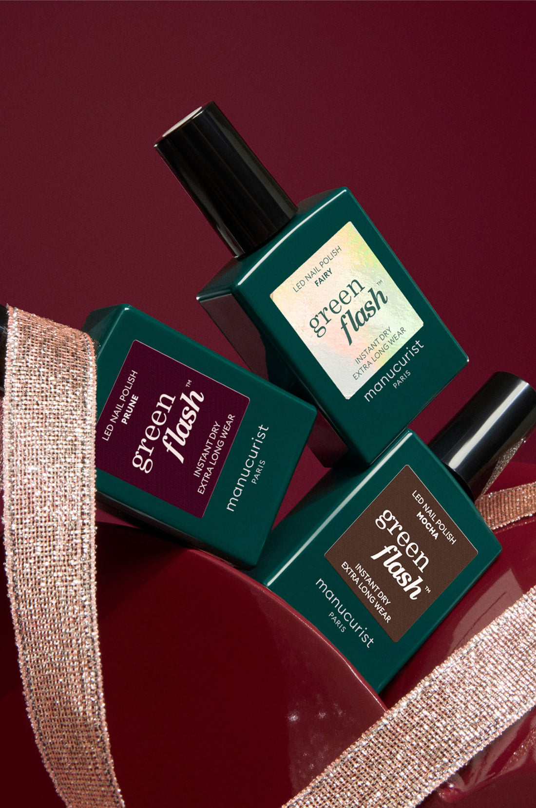 Three bottles of Manucurist Green Flash LED nail polish in different shades are arranged artistically against a deep red background, partially wrapped with a shimmery ribbon.