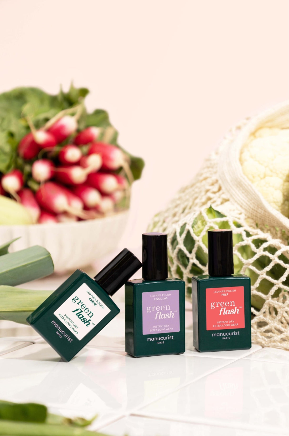 Three green bottles of Manucurist nail polish are displayed on a white surface, with radishes and a netted bag of cauliflower in the background, suggesting a fresh and natural theme.