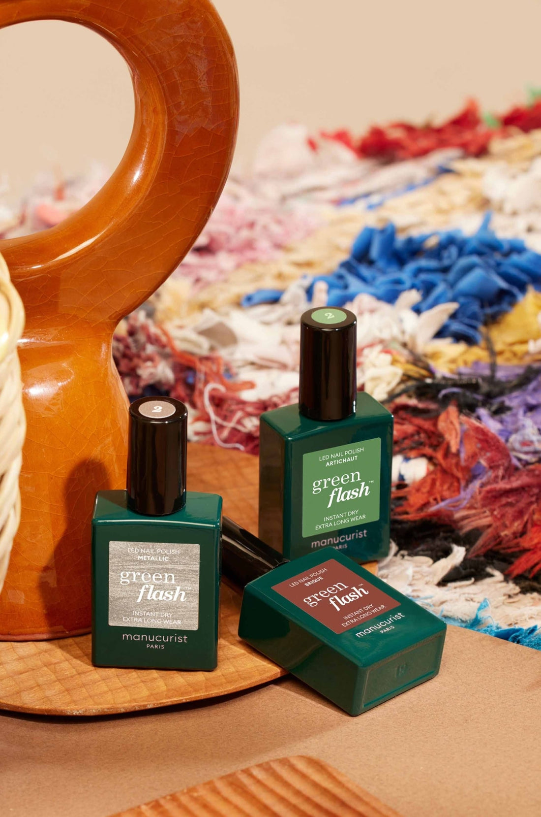 Three green bottles of Manucurist Green Flash nail polish are arranged on a wooden surface, with a textured, multicolored fabric and a brown ceramic vase in the background.