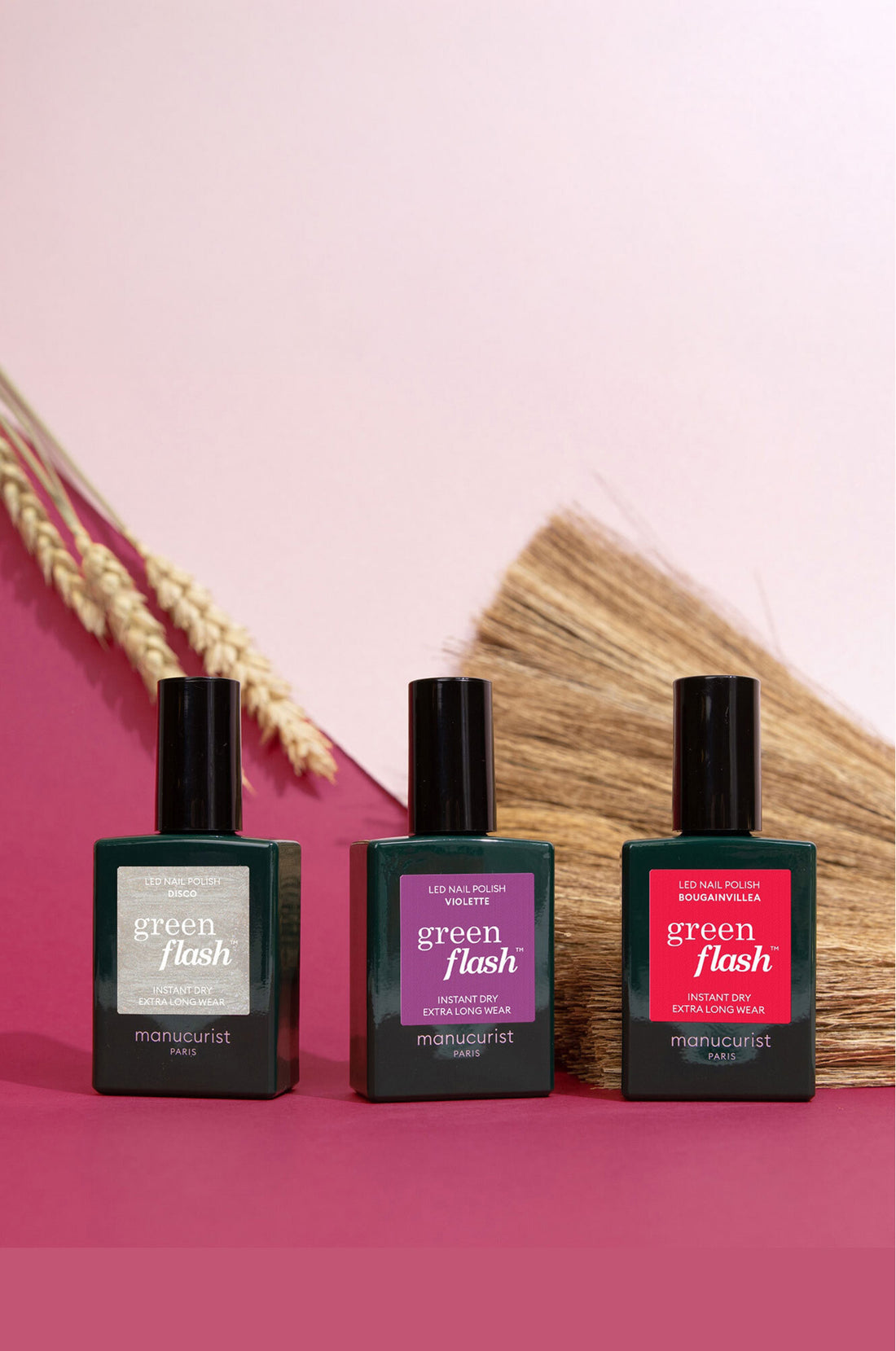 Three bottles of Manucurist Green Flash nail polish in dark green, mauve, and red shades are displayed upright on a pink surface, with dried wheat and straw in the background.