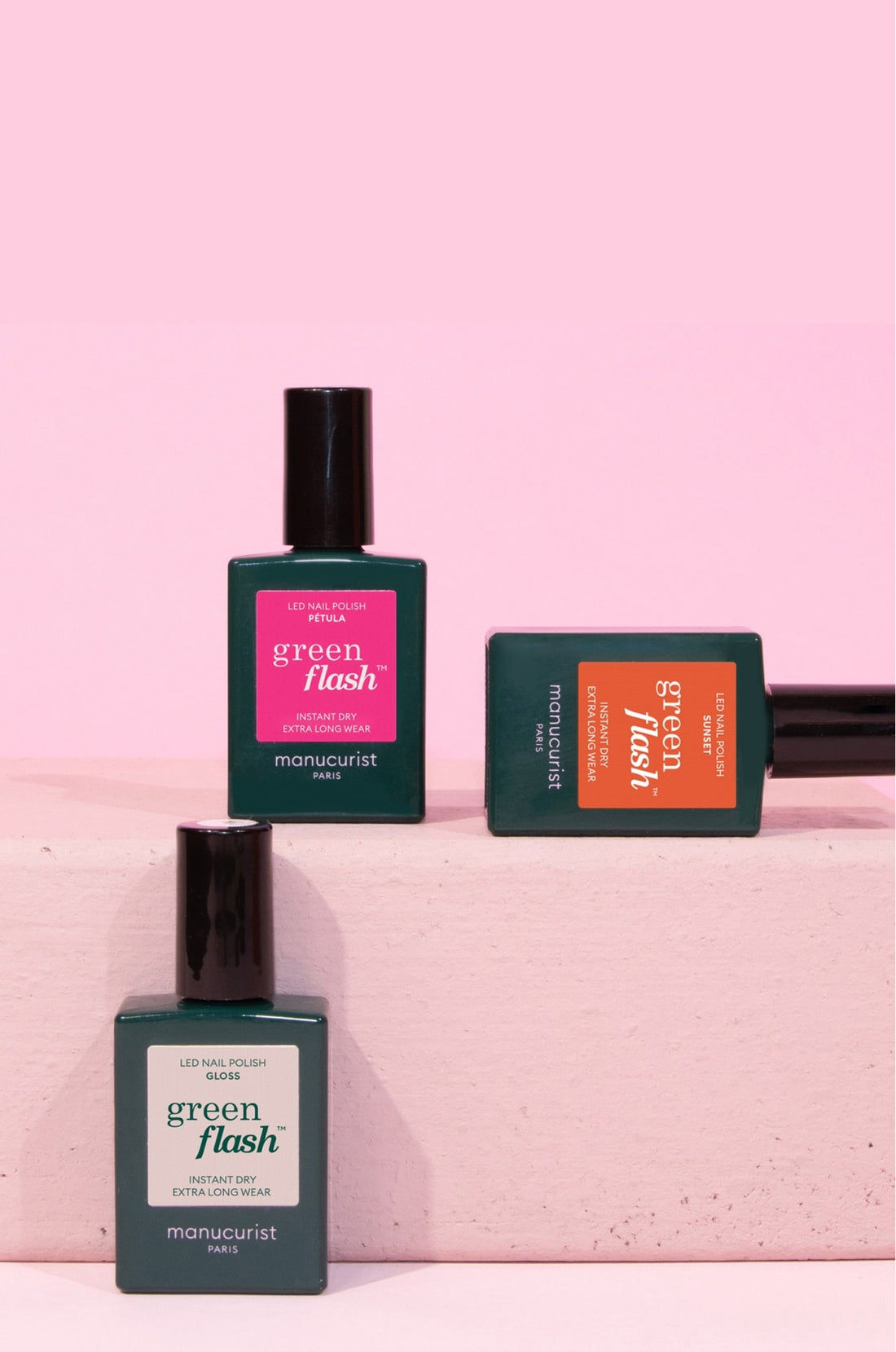 Three bottles of Green Flash LED nail polish by Manucurist, in green, pink, and orange labels, arranged on and around a pink rectangular block against a pink background.