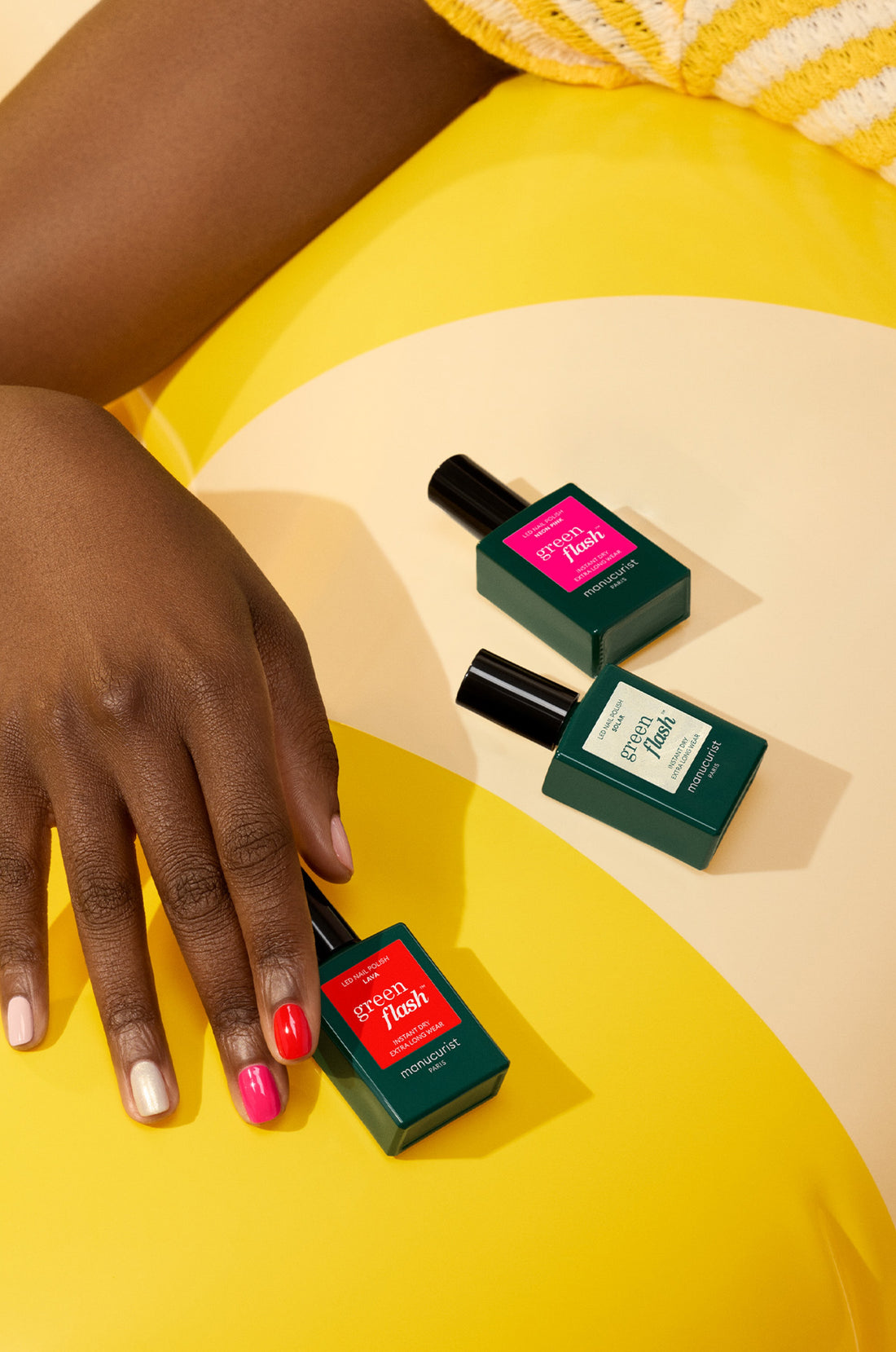 A hand with painted nails rests on a yellow surface next to three bottles of nail polish in green bottles with colorful labels, featuring shades of red, pink, and light green.