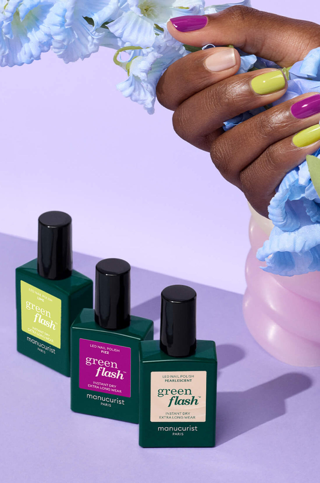 A hand with nails painted yellow, green, and purple holds light blue flowers above three bottles of Green Flash nail polish on a purple surface.