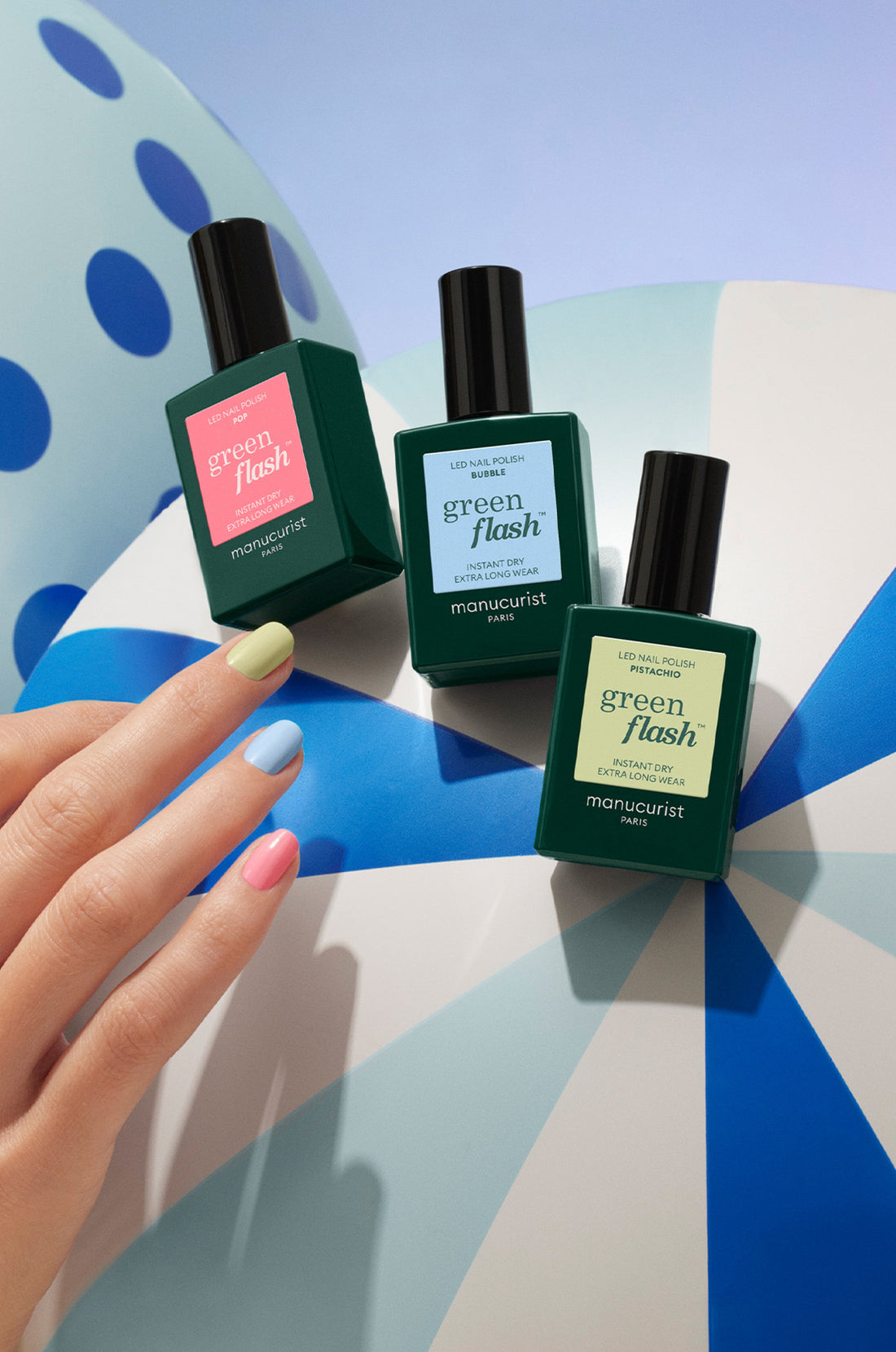 A hand with pastel-colored nails rests on a large, inflatable beach ball beside three bottles of Manucurist Green Flash nail polish in different shades.
