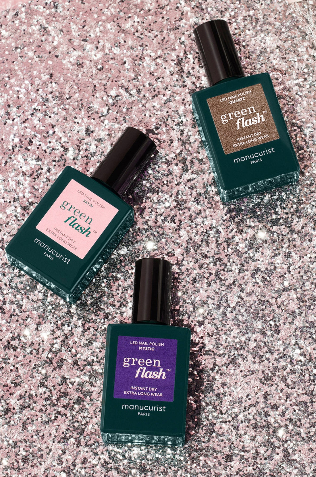 Three bottles of Manucurist Green Flash LED nail polish in pink, glittery bronze, and purple shades are arranged diagonally on a sparkling, pink glitter background.