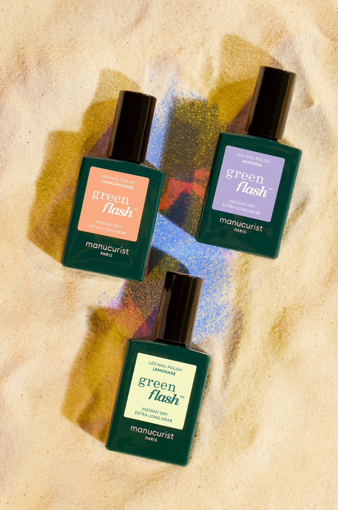 Three bottles of Green Flash nail polish by Manucurist, each with a different colored label (coral, blue, and green), are arranged on sand with colorful powder scattered around them.