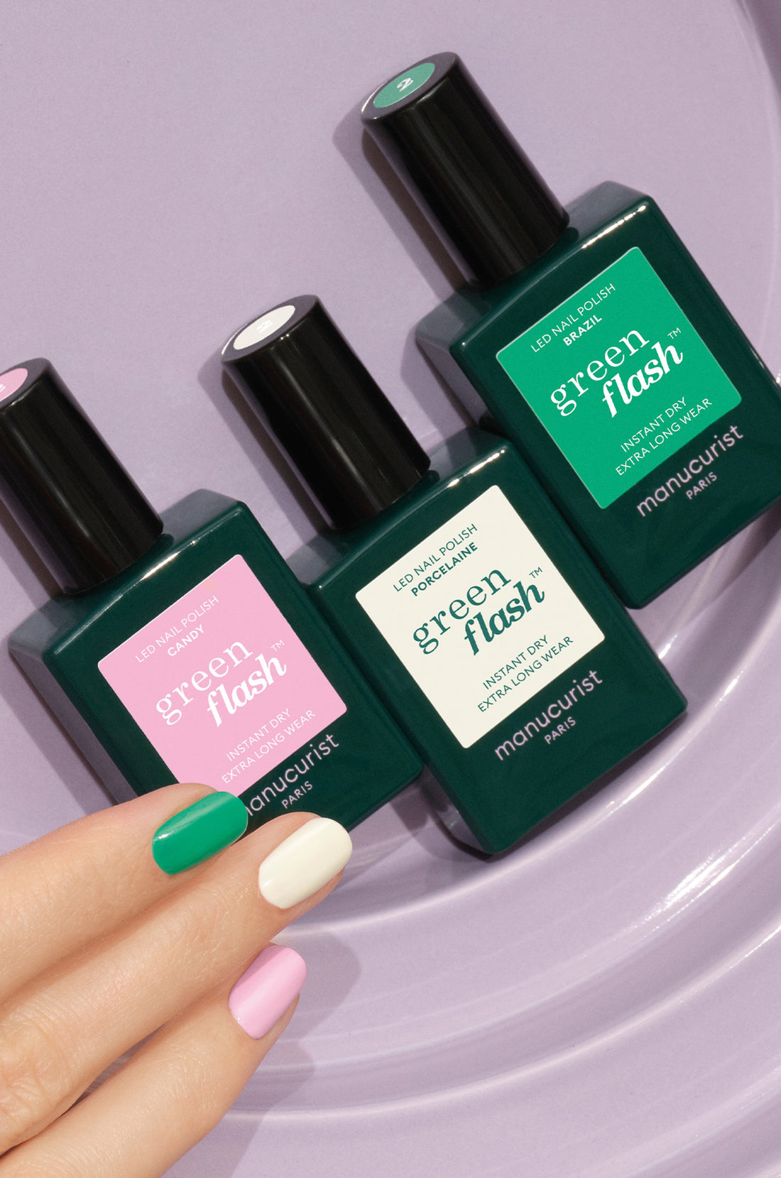 Three bottles of Manucurist Green Flash nail polish sit on a purple surface. A hand with manicured nails, each painted a different pastel shade—green, pink, cream, and lilac—rests nearby.