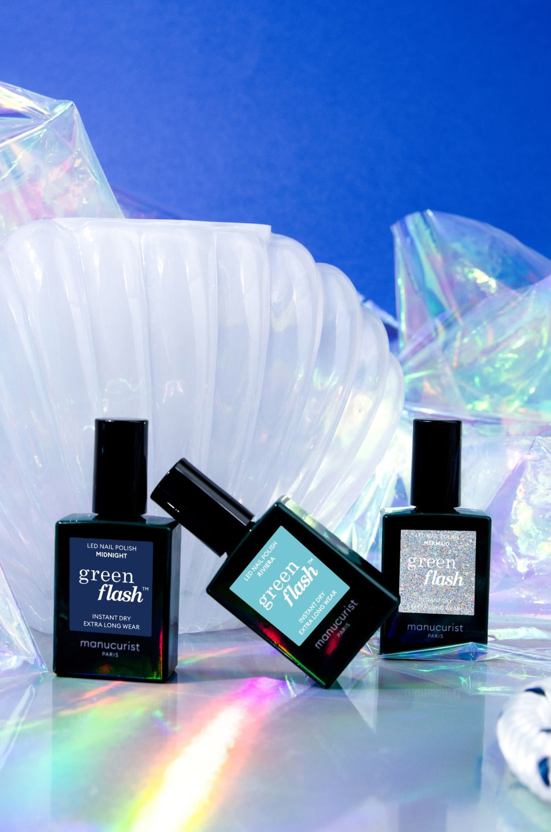 Three bottles of Manucurist Green Flash nail polish are displayed in front of an iridescent shell-like backdrop and shiny, holographic decorations against a vibrant blue background.