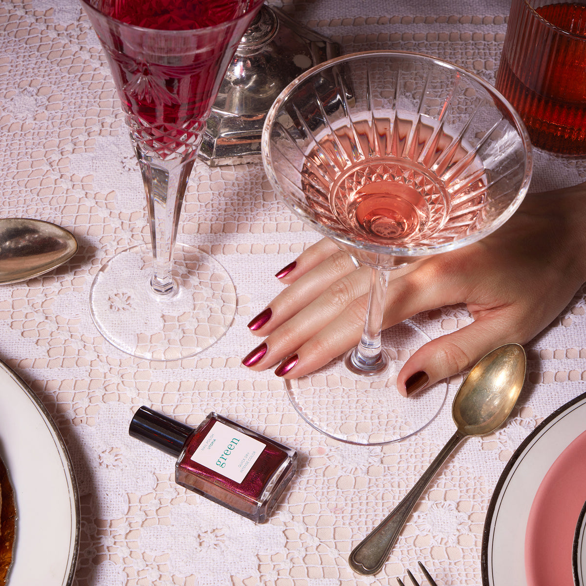 A hand with metallic crimson nails painted in Manucurist’s Utopia holds a glass of pink wine on a lace tablecloth, beside another wine glass, a Utopia bottle, silverware, and a white plate.