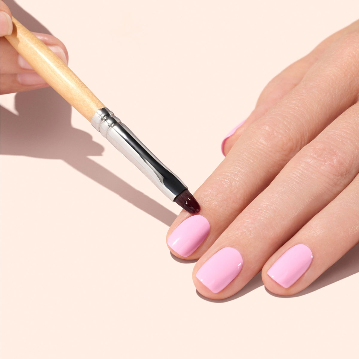 Une main aux ongles courts peints en rose clair est soigneusement nettoyée au niveau des cuticules à l’aide du Flat Brush Manucurist. L’arrière-plan est d’un beige doux.