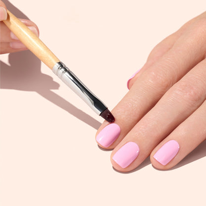 A hand with short, light pink painted nails is being carefully tidied at the cuticles using the Manucurist Flat Brush. The background is a soft beige color.