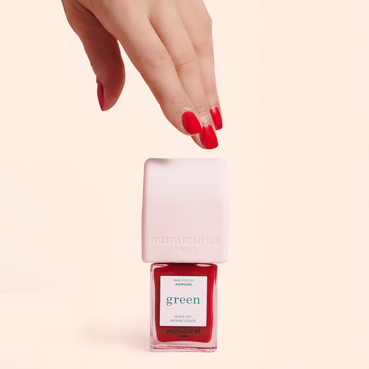 A hand with red-painted nails hovers above a bottle of Manucurist Paris green nail polish, which has a white cap and red polish inside, set against a light pink background.