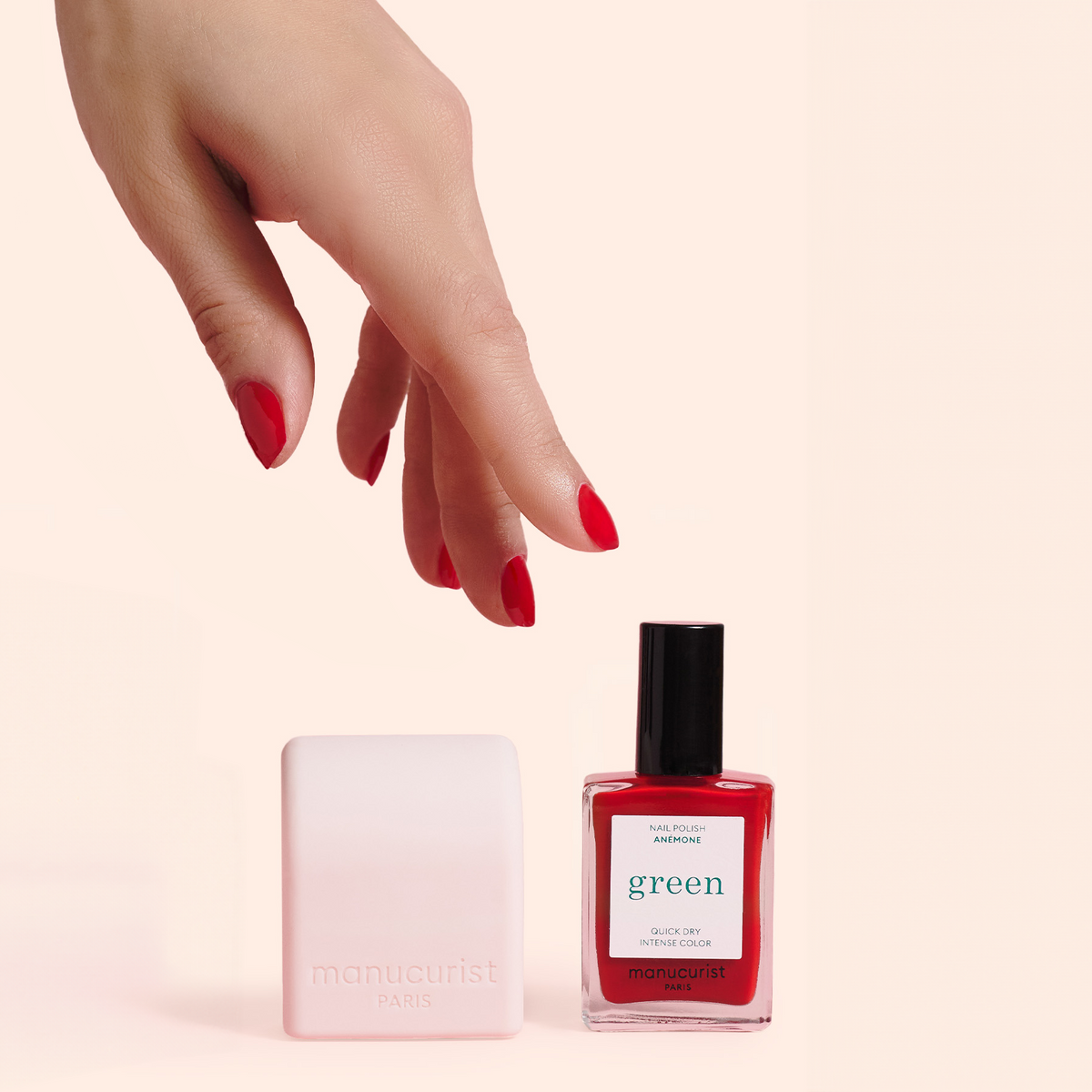 A hand with red-painted nails reaches toward a white manicure block and a bottle of red nail polish labeled green by Manucurist, set against a light pink background.